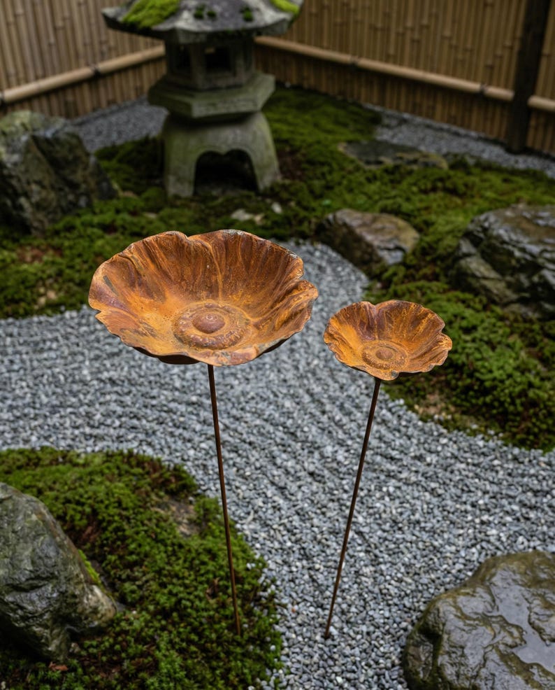 Puede incluir: Dos esculturas de flores de metal oxidado con un acabado texturizado. Las flores est&aacute;n sobre finos tallos, colocadas en un jard&iacute;n zen con grava gris y musgo verde. Una linterna de piedra y una valla de bamb&uacute; est&aacute;n en el fondo.