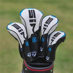 May include: A collection of golf club head covers in a golf bag. The covers are white with black and blue detailing, displaying the number "SI7" in large black lettering. The golf bag is black with a red stripe.