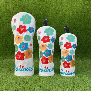 May include: Three white golf club head covers with colorful embroidered flower designs. The flowers are in shades of red, yellow, blue, and teal. The word "Flowers" is written in blue script at the bottom of each cover.