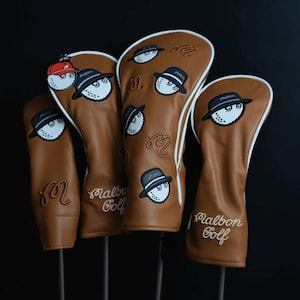 May include: Four brown golf club head covers, each with a white golf ball wearing a hat. The covers display the text "Malbon Golf" and a stylized "M" logo. They have white trim and are on black stands.