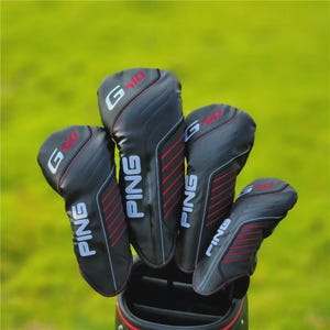 Puede incluir: Un juego de fundas negras para palos de golf con detalles en rojo y blanco. Las fundas tienen la palabra "PING" en blanco y "G410" en rojo. Las fundas están en una bolsa de golf negra sobre un fondo verde.