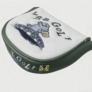 May include: White and green golf club head cover with embroidered text and graphics. The cover features the text "L.A.B. GOLF" and a stylized illustration of a bridge and a bear. The edges are green.