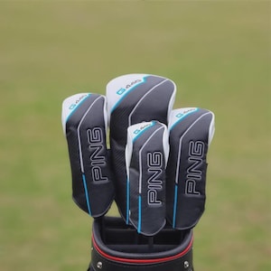 Puede incluir: Un juego de fundas para palos de golf PING en una bolsa de golf. Las fundas son negras, blancas y grises con detalles en azul. La palabra "PING" está impresa en blanco en el lateral de cada funda. El fondo es un campo verde borroso.