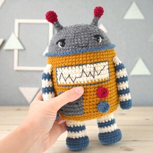 Puede incluir: Un juguete de robot de ganchillo hecho a mano en tonos grises, amarillos, azules y rojos. El robot tiene un cofre cuadrado con un diseño en zigzag, brazos y piernas a rayas, y dos antenas rojas. El juguete es sostenido por una mano.