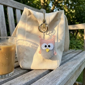 May include: A cream-colored canvas tote bag with a brass zipper and a crocheted owl keychain. The owl is gray, pink, and yellow. A glass of iced coffee sits to the left of the bag on a wooden bench.
