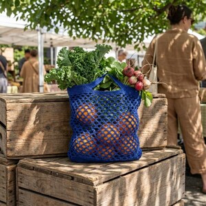 Op de afbeelding: Een blauwe gehaakte herbruikbare boodschappentas gevuld met sinaasappels, boerenkool en radijsjes. De tas staat op een stapel houten kisten op een markt in de open lucht. Een persoon in een bruin outfit staat op de achtergrond.