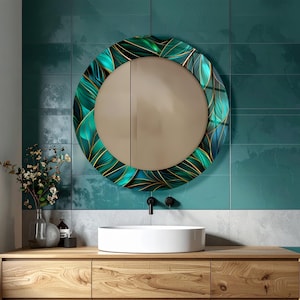 May include: A round bathroom mirror with a decorative leaf design in shades of green and gold. The mirror is mounted on a teal tiled wall above a wooden vanity with a white sink and black faucet.