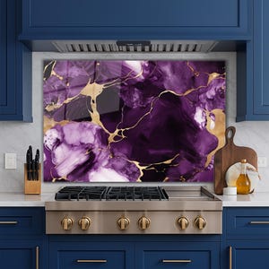 May include: A decorative kitchen splashback with a purple and gold marble effect. The design features swirling shades of purple with gold veining. The splashback is installed behind a stainless steel cooker and is a decorative element.