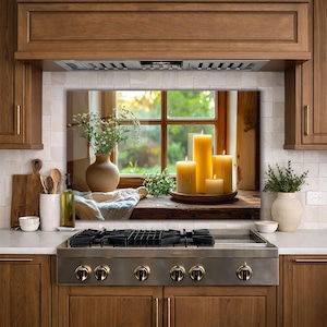 May include: A kitchen scene featuring a window with lit candles on a wooden windowsill. A vase with flowers and a small plant are also on the sill. The candles are yellow and of varying heights. The kitchen has wooden cabinets and a stainless steel stove.