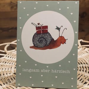 Könnte beinhalten: Grußkarte mit einer Aquarellillustration einer Schnecke, die ein Geschenk trägt. Die Schnecke hat ein graues Gehäuse mit weißen Punkten und einen roten Körper. Der Hintergrund ist hellgrün mit weißen Herzen, und der Text "langsam aber härzlech" steht unten.