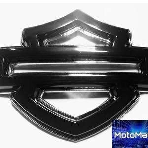 Puede incluir: Un emblema negro brillante con forma de escudo, con una barra horizontal y una forma superior e inferior estilizada. El emblema tiene una superficie reflectante y está sobre un fondo blanco. El logotipo de MotoMatrix está en la esquina inferior derecha.