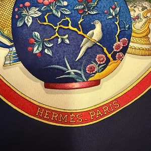 May include: A silk scarf with a detailed illustration. The design features a blue vase with a bird perched on a branch with pink flowers and red berries. The scarf has a red border with "HERMES-PARIS" printed in gold.