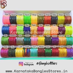 Puede incluir: Una exhibición de pulseras coloridas en varios tonos, incluyendo rosa, verde y dorado. Las pulseras están dispuestas en filas y etiquetadas con nombres de colores. El texto "Karnataka Bangles Store" es visible.