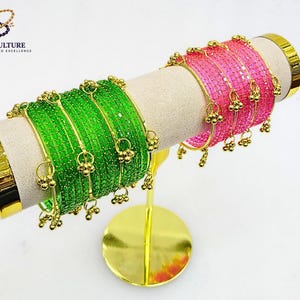 Könnte beinhalten: Grüne und rosa Armreifen mit goldenen Details werden auf einem goldenen Ständer präsentiert. Die Armreifen sind mit kleinen goldenen Anhängern verziert. Der Markenname "BANGLES CULTURE" ist oben links sichtbar.