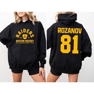 May include: Black hoodie with the text "RAIDERS BOSTON HOCKEY" in gold lettering on the front. The back of the hoodie displays the name "ROZANOV" and the number "81" in gold. The hoodie has a front pocket and a hood.
