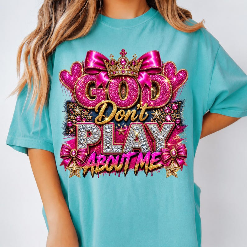 May include: A turquoise t-shirt featuring a graphic design with the text "GOD Don't PLAY ABOUT ME" in glittery pink and diamond-textured letters. The design includes a gold crown, pink bows, hearts, and gold stars on a dark background.