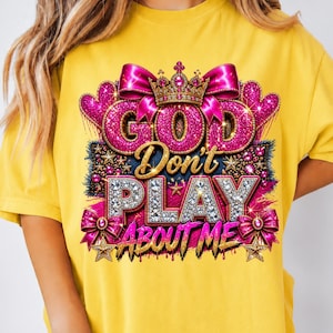 May include: A bright yellow t-shirt featuring a graphic design with the text "GOD Don't PLAY ABOUT ME". The design includes a gold crown, pink glittery hearts, bows, and stars, with the word "GOD" in pink glitter and "PLAY" in silver rhinestones.