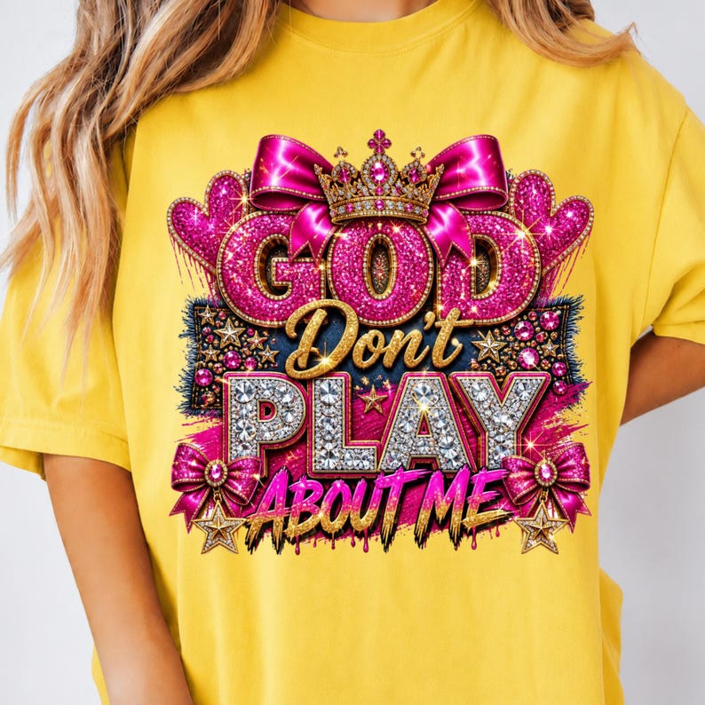 May include: A bright yellow t-shirt featuring a graphic design with the text "GOD Don't PLAY ABOUT ME". The design includes a gold crown, pink glittery hearts, bows, and stars, with the word "GOD" in pink glitter and "PLAY" in silver rhinestones.