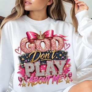 May include: A white long-sleeve sweatshirt featuring a graphic print with the text "GOD Don't PLAY ABOUT ME". The design includes pink glitter lettering, a gold crown, a pink satin bow, denim-textured accents, and small star embellishments.