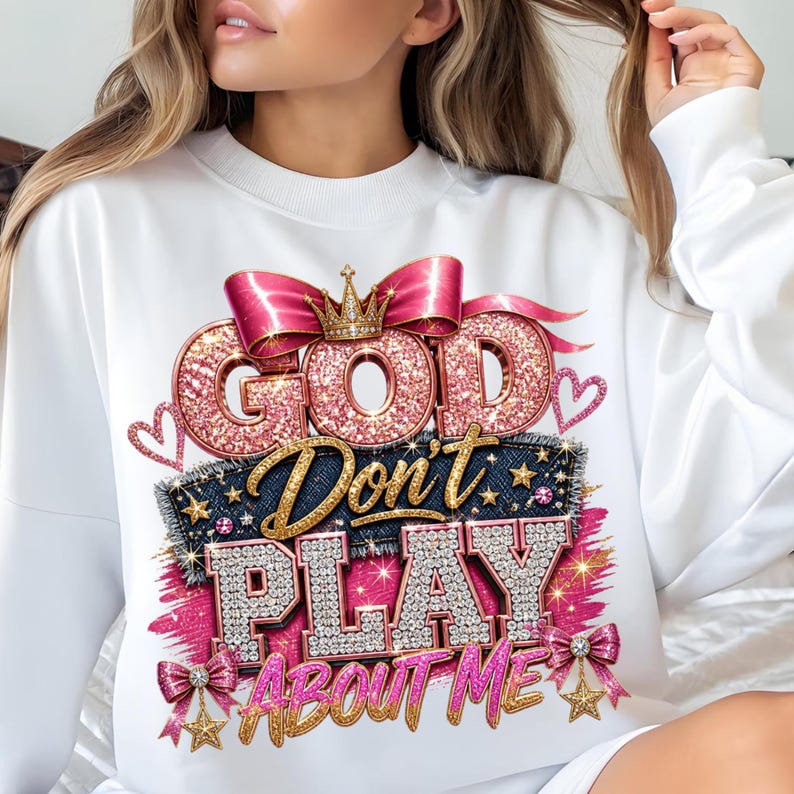 May include: A white long-sleeve sweatshirt featuring a graphic print with the text "GOD Don't PLAY ABOUT ME". The design includes pink glitter lettering, a gold crown, a pink satin bow, denim-textured accents, and small star embellishments.
