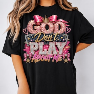 May include: A black t-shirt featuring a graphic design with the text "GOD Don't PLAY ABOUT ME". The design includes pink glittery lettering, a gold crown, pink bows, and diamond-textured accents, all arranged in a layered, decorative style.