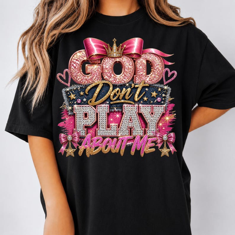 May include: A black t-shirt featuring a graphic design with the text "GOD Don't PLAY ABOUT ME". The design includes pink glittery lettering, a gold crown, pink bows, and diamond-textured accents, all arranged in a layered, decorative style.