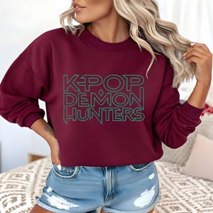 May include: A maroon sweatshirt with the text "K-POP DEMON HUNTERS" in green block letters. The sweatshirt has long sleeves and a crew neck. The model is wearing blue denim shorts.