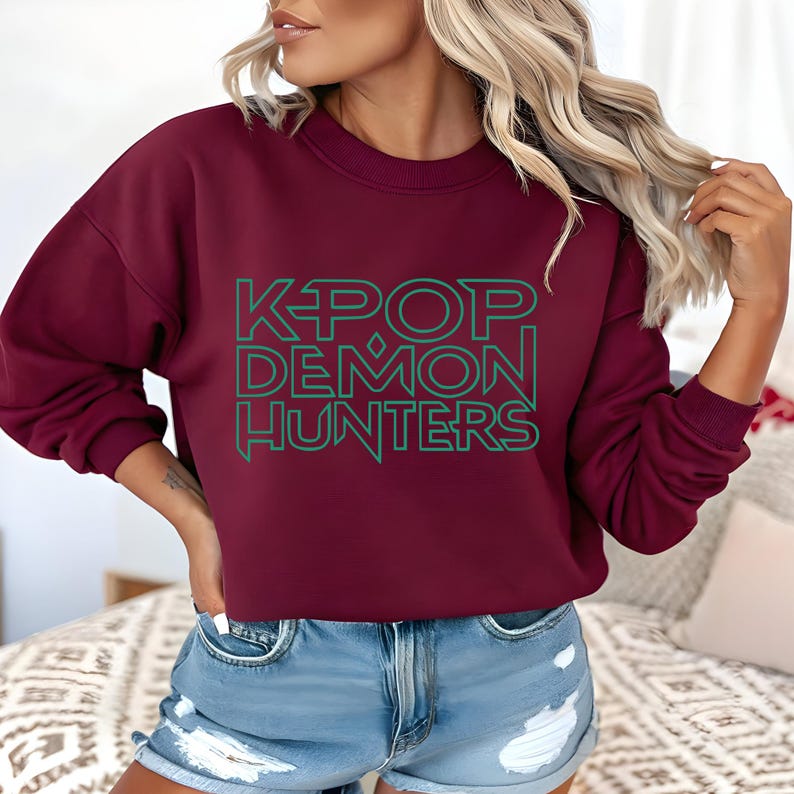 May include: A maroon sweatshirt with the text "K-POP DEMON HUNTERS" in green block letters. The sweatshirt has long sleeves and a crew neck. The model is wearing blue denim shorts.