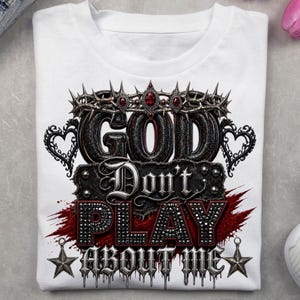 May include: A white t-shirt featuring a graphic design with the text "GOD Don't PLAY ABOUT ME". The design includes a crown of thorns with red gems, heart-shaped filigree, a red paint splatter effect, and two five-pointed stars at the bottom.