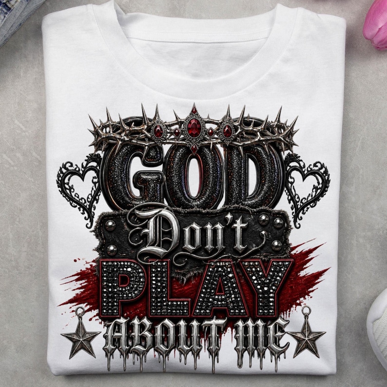 May include: A white t-shirt featuring a graphic design with the text "GOD Don't PLAY ABOUT ME". The design includes a crown of thorns with red gems, heart-shaped filigree, a red paint splatter effect, and two five-pointed stars at the bottom.