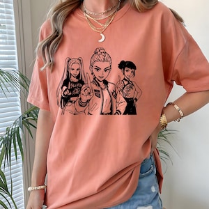 May include: A peach-colored t-shirt featuring a black and white graphic of three anime-style women. The central figure points upwards, while the others gesture. One shirt has the word "SLAYER" printed on it.