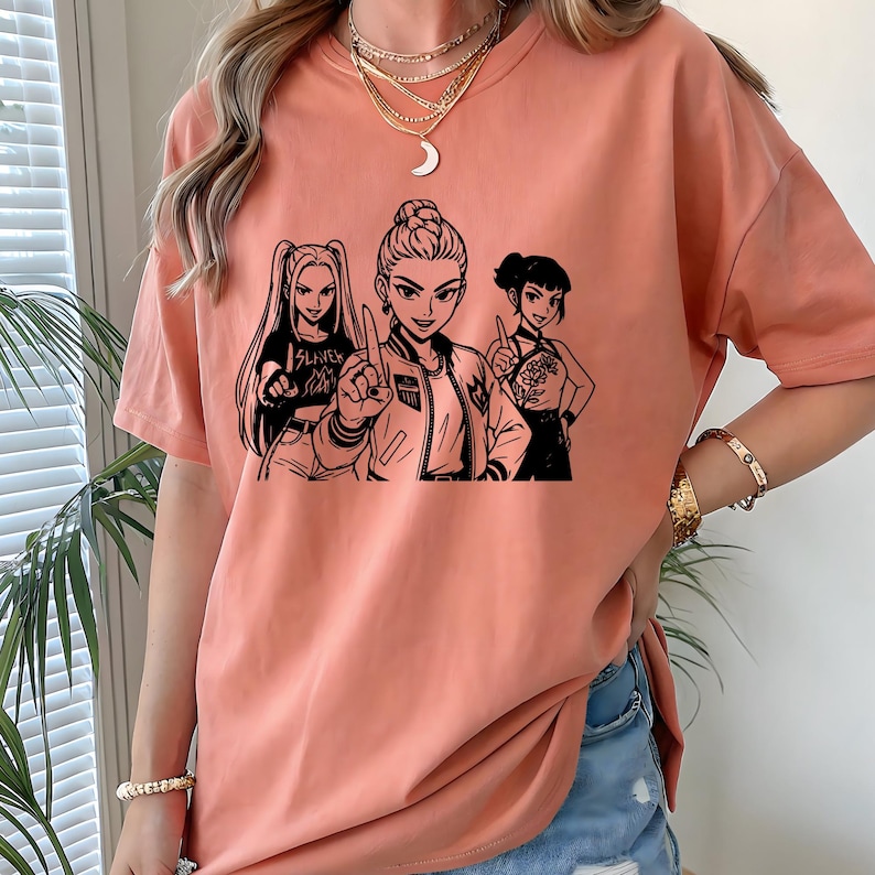 May include: A peach-colored t-shirt featuring a black and white graphic of three anime-style women. The central figure points upwards, while the others gesture. One shirt has the word "SLAYER" printed on it.