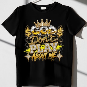 May include: A black t-shirt featuring a graphic design with a gold crown above the text "GOD Don't PLAY ABOUT ME" in metallic and diamond-like lettering. The design includes dollar signs, flames, and star-shaped accents in gold and silver.