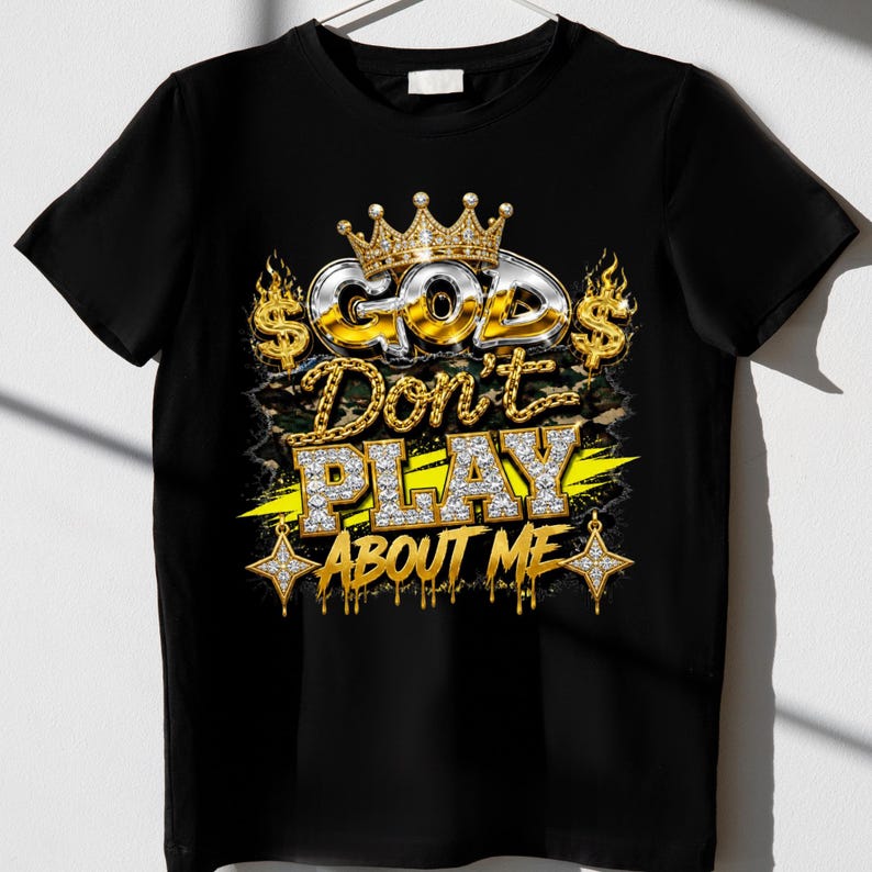 May include: A black t-shirt featuring a graphic design with a gold crown above the text "GOD Don't PLAY ABOUT ME" in metallic and diamond-like lettering. The design includes dollar signs, flames, and star-shaped accents in gold and silver.