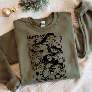 May include: An olive green sweatshirt with a black graphic design featuring three anime-style characters and the text "HUNTR/X". The names "Mira", "Rumi", and "Zoey" are also included. The sweatshirt is folded on a white surface.