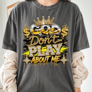May include: A charcoal gray t-shirt featuring a graphic print with the text "GOD Don't PLAY ABOUT ME". The design includes a gold crown, dollar signs, chain links, and diamond-textured lettering over a camouflage background with yellow accents.
