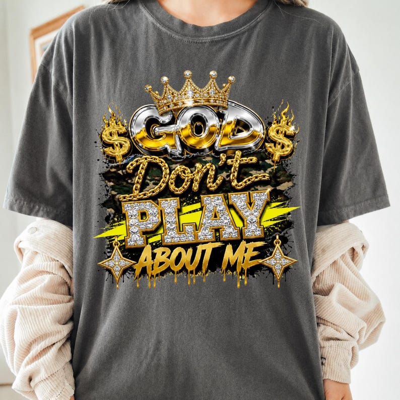 May include: A charcoal gray t-shirt featuring a graphic print with the text "GOD Don't PLAY ABOUT ME". The design includes a gold crown, dollar signs, chain links, and diamond-textured lettering over a camouflage background with yellow accents.