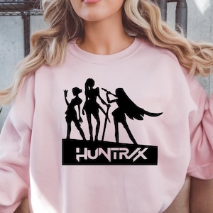 May include: A light pink sweatshirt featuring a black graphic of three stylized figures with weapons, standing on a black rectangle with the word "HUNTRX" in white lettering. The sweatshirt has long sleeves.