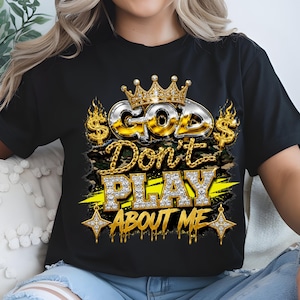 May include: A black t-shirt featuring a graphic design with the text "GOD Don't PLAY ABOUT ME" in metallic and diamond-textured lettering. The design includes a gold crown, dollar signs, flames, and star accents over a camouflage background.