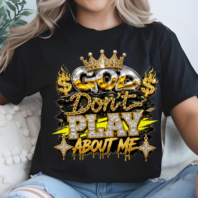 May include: A black t-shirt featuring a graphic design with the text "GOD Don't PLAY ABOUT ME" in metallic and diamond-textured lettering. The design includes a gold crown, dollar signs, flames, and star accents over a camouflage background.