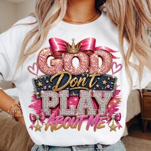 May include: A white crewneck sweatshirt featuring a graphic print with the text GOD DON'T PLAY ABOUT ME. The design includes pink glittery letters, a gold crown, pink bows, denim-textured accents, and gold stars on a bright pink brushstroke.