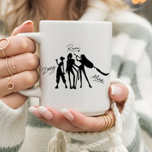 May include: White ceramic mug featuring a black silhouette design of three figures with the names Zoey, Rumi, and Mira. The mug has a large handle and is held by a person wearing a striped sweater.