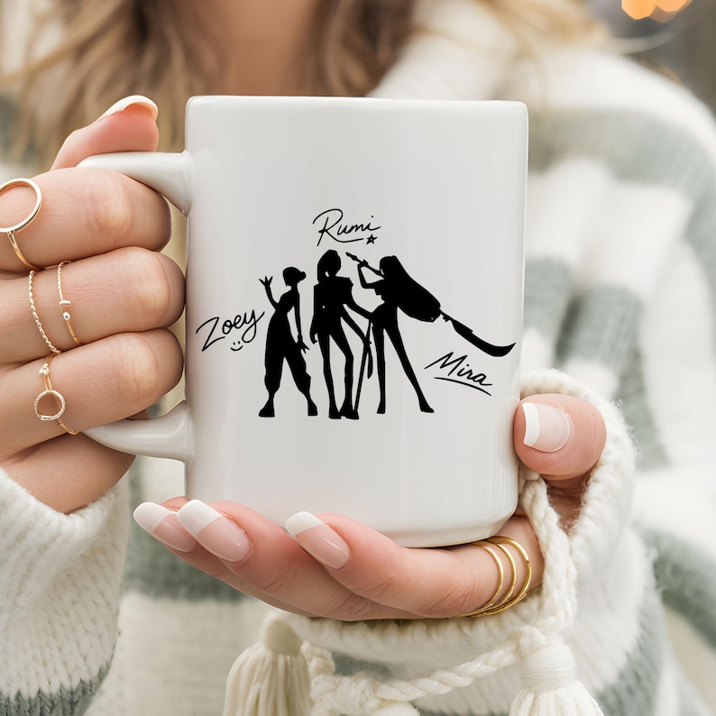 May include: White ceramic mug featuring a black silhouette design of three figures with the names Zoey, Rumi, and Mira. The mug has a large handle and is held by a person wearing a striped sweater.