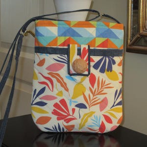 May include: A white crossbody bag with a colorful geometric pattern on the top flap and a floral pattern on the bottom. The bag has a blue denim strap and a brown button closure.