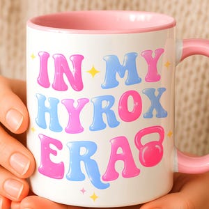 May include: A white ceramic mug with a pink interior and handle, featuring the text "IN MY HYROX ERA" in a bubbly, retro-style font with alternating pink and blue letters, accompanied by small yellow star accents and a pink kettlebell icon.