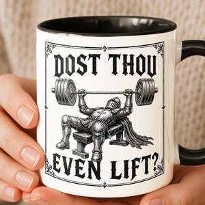 Pode incluir: Caneca de cerâmica branca com interior e alça pretos. A caneca apresenta um gráfico de um cavaleiro levantando pesos com o texto "DOST THOU EVEN LIFT?" em uma fonte decorativa. O design é em preto e branco.