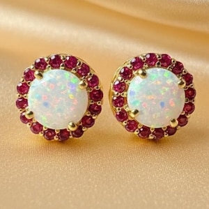 May include: A pair of gold-colored stud earrings. Each earring features a large, round, white opal center stone surrounded by a halo of small, red gemstones. The earrings are set against a gold-colored, satin-like background.