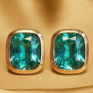 May include: A pair of gold-toned square stud earrings. Each earring features a large, emerald-green gemstone with a faceted cut. The gemstones are set in a gold bezel setting, creating a luxurious look. The background is a solid, golden-yellow color.