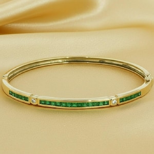 Könnte beinhalten: Ein goldfarbenes Armband mit rechteckigen smaragdgrünen Steinen und kleinen klaren Steinen. Das Armband ist vor einem goldfarbenen, satinartigen Hintergrund platziert.