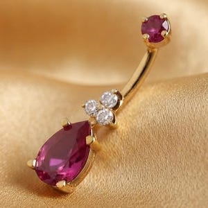 May include: A gold navel piercing ring featuring a curved barbell with a round, deep pink gemstone at the top. The bottom end displays a teardrop-shaped deep pink gemstone accented by three small, clear, round white stones set in a cluster.